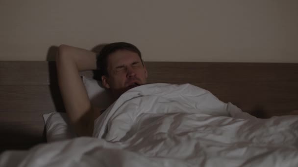 Ung mand ligger på pude under tæppe i sengen og ser tv i slutningen af aftenen. Afslappet mand gaber og falder i søvn, mens du ser tv i mørkt rum. – Stock-video
