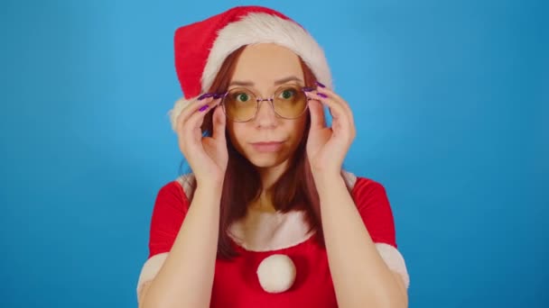 サンタの衣装を着た女性は 青い背景に立って 彼女の眼鏡を調整します クリスマスの帽子の女性は カメラを見て 彼女の顔に眼鏡を調整します — ストック動画