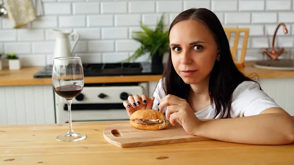 年轻女人吃汉堡包 坐在厨房的桌子边 布伦内特看着镜头 一边吃着东西 一边喝酒 — 图库照片