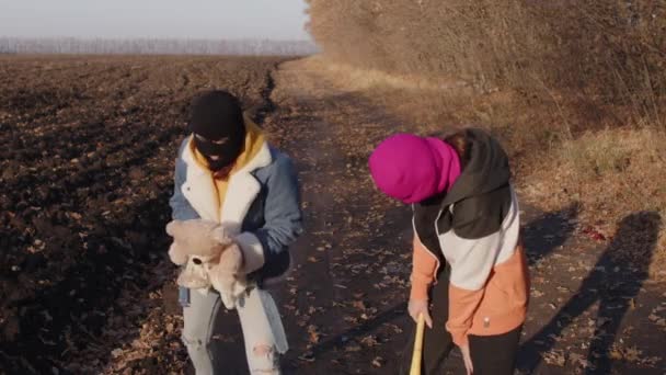 Women in balaclava with baseball bat and teddy bear in countryside. One female swings bat to plush toy of girlfriend. — Stock Video