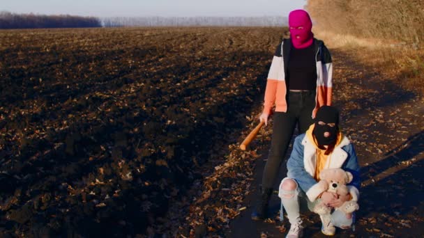 Kvinnor i Balaclava med baseballträ och nallebjörn på landet. En kvinna står med fladdermus, andra huk nära flickvän med plysch leksak i händerna. — Stockvideo