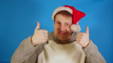 Noel Baba şapkalı adam mavi arka planda baş parmak gösteriyor. Noel şapkalı bir erkek gülümsüyor ve kameraya bakıyor..