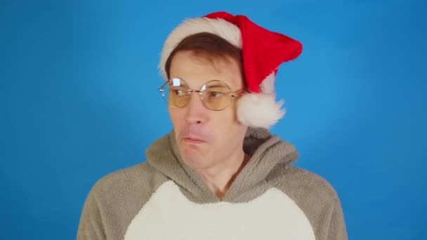 サンタの帽子と眼鏡の男は青い背景に舌を示しています 男性でクリスマス帽子スティックアウト舌とそれを隠します イチャイチャ — ストック動画