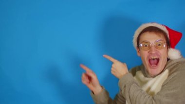 Noel Baba şapkalı ve gözlüklü adam mavi arka planda mesaj için yer gösteriyor. Yılbaşı şapkalı bir erkek yeni yıl satışlarıyla şoke oldu..