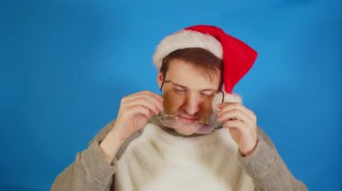 Noel Baba şapkalı adam mavi arka planda duran gözlük takıyor. Noel şapkalı ve gözlüklü bir adam kameraya bakıyor..