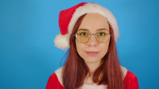 Woman in santa costume and glasses looking at camera and smiling. Female in christmas hat and eyeglasses laughs, standing on blue background. — Stock Video