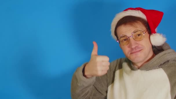 サンタの帽子と眼鏡の男は青い背景のテキストのための指のスペースを指します。新年の販売によって楽しくショックを受けたクリスマスの帽子の男性. — ストック動画