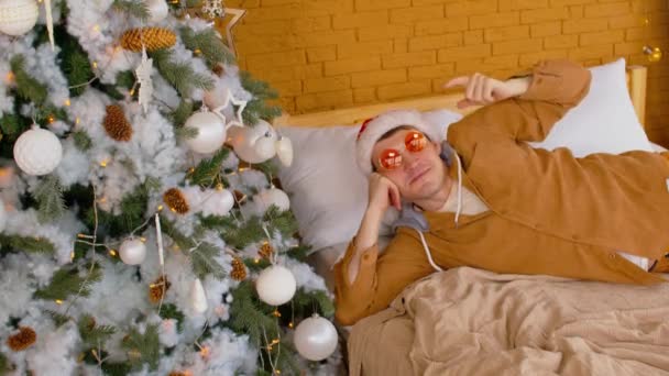 Homem de chapéu de Papai Noel e óculos de sol deitado na cama perto da árvore de Natal decorada. Homem elegante aponta o dedo para a árvore de coníferas com diferentes brinquedos de Natal, olhando para a câmera e sorrindo. — Vídeo de Stock
