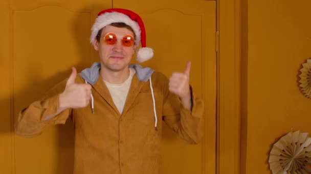 サンタの帽子とサングラスの男は、部屋の茶色のドアの背景に立って、親指を表示します。スタイリッシュな男性の笑顔とカメラを見て、承認のジェスチャーを示す. — ストック動画
