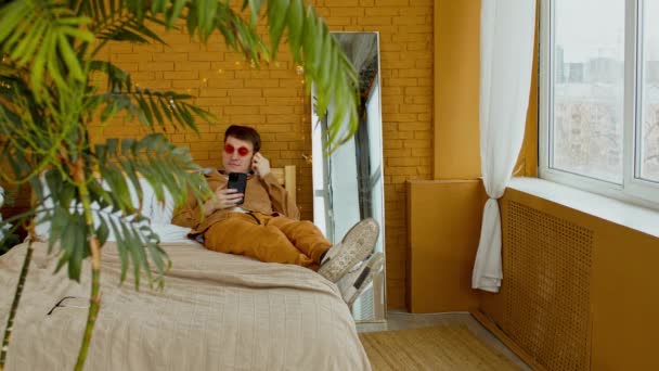 Muž ve slunečních brýlích ležel na posteli s mobilním telefonem a šťoural se v uchu. Stylový mužský odpočinek a procházení smartphone uvnitř. — Stock video