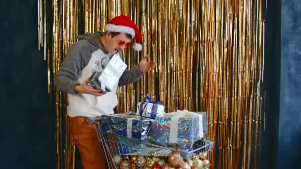Giovane maschio in occhiali luminosi e cappello di Babbo Natale in piedi con carrello pieno di bagattelle assortite e avvolto regali di Natale contro parete con fili scintillanti. — Video Stock