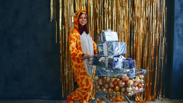 Mulher sorridente em kigurumi pijama de pé com carrinho com montão de várias bugigangas decorativas e caixas de Natal perto da parede com ouropel dourado — Vídeo de Stock