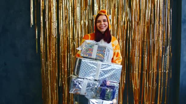 Ευτυχισμένη γυναίκα με τρόλεϊ με χριστουγεννιάτικα δώρα και μπιχλιμπίδια. Χαμογελαστή γυναίκα σε kigurumi πυτζάμα στέκεται με καλάθι με σωρό διακοσμητικά διάφορα μπιχλιμπίδια — Αρχείο Βίντεο