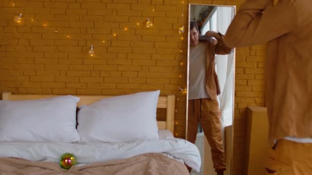 Visszapillantás a termés férfi néz tükörbe, és felvesz sárga ing az ágy mellett diszkó labda takaró a hálószobában díszített tündér fények karácsonyra — Stock videók