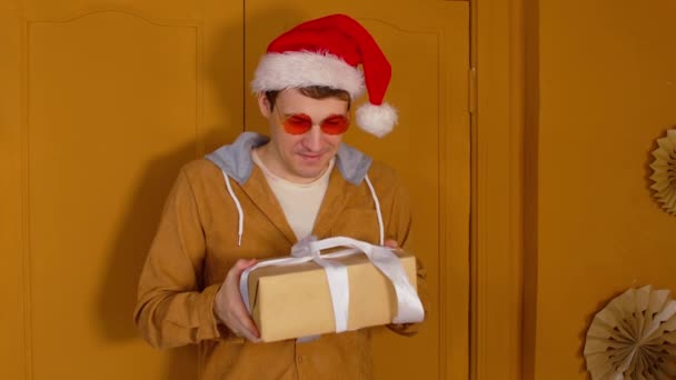 Chapeau homme au Père Noël avec cadeau de Noël en boîte. Enfant mâle dans des lunettes de soleil lumineuses et chapeau de Père Noël debout avec boîte cadeau de Noël attaché avec ruban dans la chambre avec des murs jaunes — Video