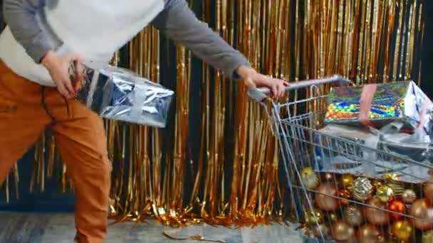 Onherkenbaar persoon met winkelwagentje gevuld met diverse kerstballen en geschenkdozen. Onbekende man rolt kar met gewikkelde kerstcadeaus, kerstversieringen en zet een ander cadeau in trolley. — Stockvideo