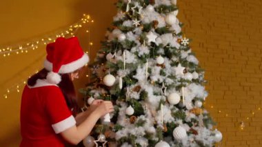 Noel Baba kostümlü bir kadının Noel ağacı süslemesi. Noel şapkalı bir kadın Noel oyuncağını asar ve kapalı mekanlarda bayram havası yaratır..