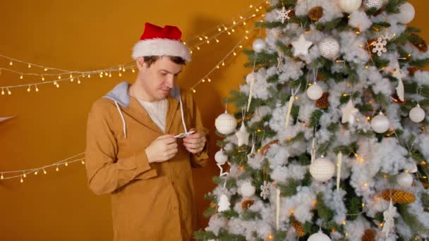 Hombre con sombrero de santa adornando el árbol de Navidad. Joven macho en sombrero de Navidad considera y cuelga juguete de Navidad, crea ambiente festivo durante las vacaciones en el interior. — Vídeos de Stock