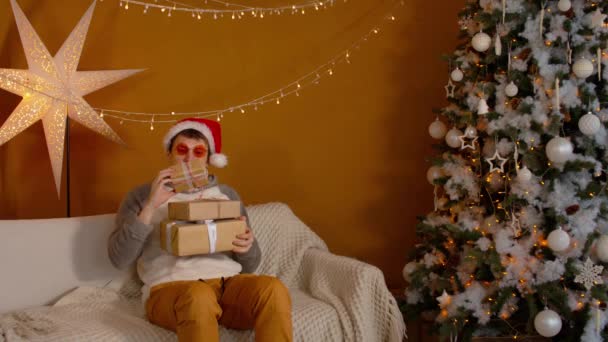 Мужчина в шляпе Санты и ярких очках с подарочными коробками, сидящими на диване возле елки. Молодой мужчина рассматривает и выбирает рождественские подарки, которые хочет открыть. Концепция сюрпризов во время праздников. — стоковое видео