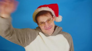 Mavi arka planda Noel Baba şapkalı ve gözlüklü mutlu adam. Genç erkek kameraya bakıyor, gülümsüyor ve el sallıyor, birine sesleniyor..