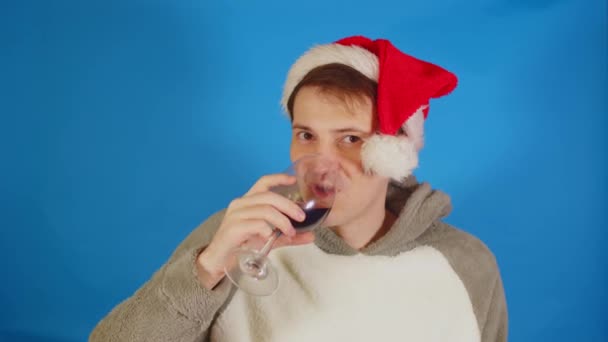 Un om fericit în pălărie de Moş Crăciun cu alcool pe fundal albastru. Un tânăr se uită la cameră, bea vin roșu și zâmbește. Conceptul de sărbătoare. — Videoclip de stoc