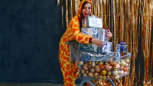 Счастливая женщина с тележкой с рождественскими подарками и безделушками. Улыбающаяся женщина в пижаме кигуруми, стоящая с тележкой с кучей различных декоративных безделушек — стоковое видео