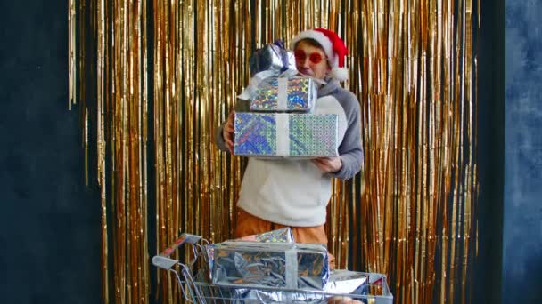 Homme excité avec chariot de boîtes-cadeaux et de boules. Jeune homme dans des lunettes lumineuses et chapeau de Père Noël debout avec chariot rempli de boules assorties et enveloppé cadeaux de Noël — Video