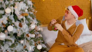 Noel Baba şapkalı ve şık gözlüklü bir adam yatakta oturuyor ve Noel ağacını süslüyor. Evde Noel kutlaması için hazırlanırken köknar dalına parlak mücevherler asan genç bir erkek..