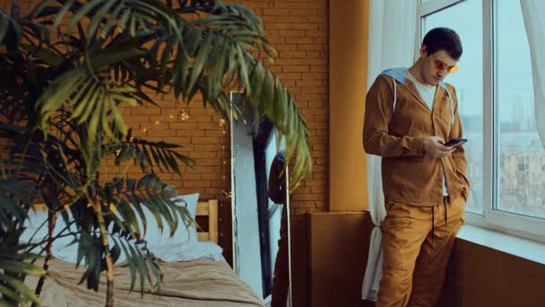 Geconcentreerde jonge man in heldere stijlvolle glazen staan in de buurt van raam en sms 'en op mobiele telefoon in de slaapkamer met palm en decoratieve gloeiende slinger voor kerstvakantie — Stockvideo