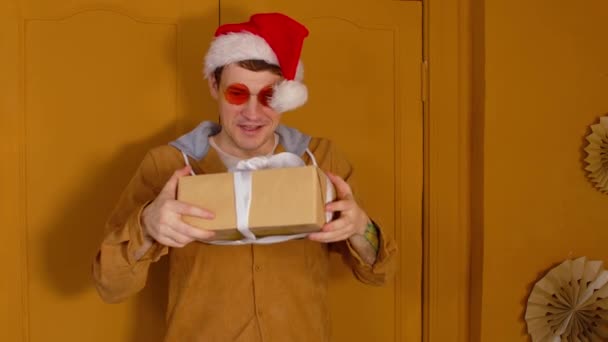 Chapeau homme au Père Noël avec cadeau de Noël en boîte. Enfant mâle dans des lunettes de soleil lumineuses et chapeau de Père Noël debout avec boîte cadeau de Noël attaché avec ruban dans la chambre avec des murs jaunes — Video