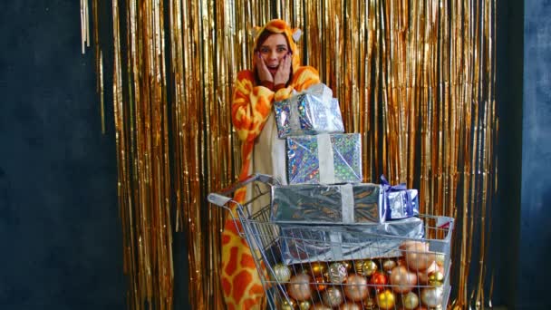 Улыбающаяся женщина в пижаме кигуруми стоит с телегой с кучей декоративных различных безделушек и рождественских коробок возле стены с золотой мишурой — стоковое видео
