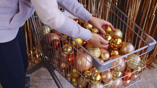 Widok z boku ziarna anonimowy kobieta wybierając złote bombki dekoracyjne w wózku do przygotowania Bożego Narodzenia stojąc przy ścianie ze złotym świecidełkiem — Wideo stockowe