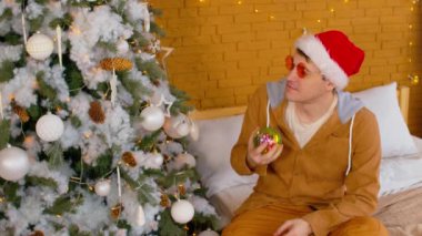 Noel Baba şapkalı ve parlak gözlüklü bir adamın portresi. Noel ağacının yanında, yatağında disko takısı takıyor. Genç erkek, süslenmiş ağaca bakıyor, düşünüyor ve elindeki Noel oyuncağını kusuyor..