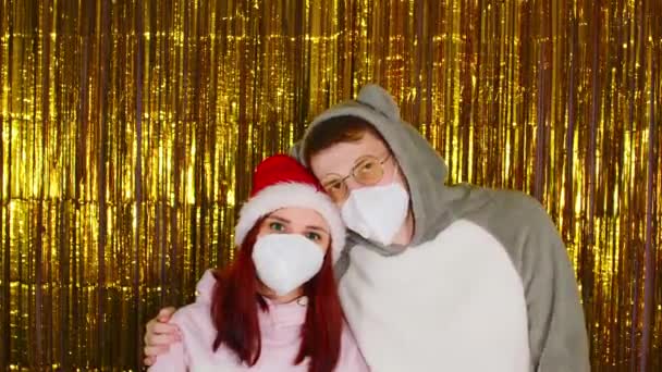 Mujer y hombre con máscaras médicas sobre fondo de oropel dorado brillante. Hombre y mujer en máscaras protectoras mirando a la cámara. Concepto de celebración de una Navidad segura durante la pandemia de coronavirus. — Vídeos de Stock