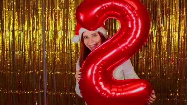 Woman in santa hat with red figure two on background of shimmering golden tinsel. Pretty female looking at camera and standing with mouth wide open. — Stock Video