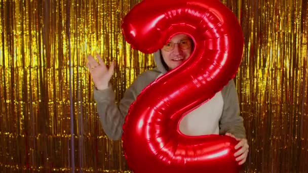Man with red figure two on background of shimmering golden tinsel. Handsome male in glasses looking at camera, smiling through numeral figure and waving hand. — Stock Video