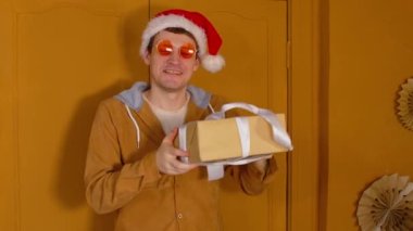 Noel Baba şapkalı, kutusunda Noel hediyesi olan bir adam. Parlak güneş gözlüklü ve Noel Baba şapkalı çocuksu bir erkek. Elinde sarı duvarlı bir kurdeleyle Noel hediyesi kutusu var.