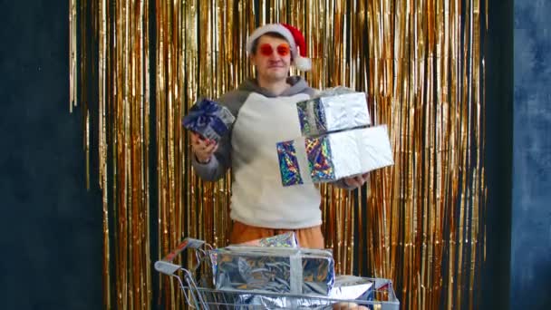 Hombre excitado con carro de cajas de regalo y adornos. Joven macho en gafas brillantes y sombrero de Santa con carro lleno de adornos surtidos y regalos de Navidad envueltos — Vídeos de Stock