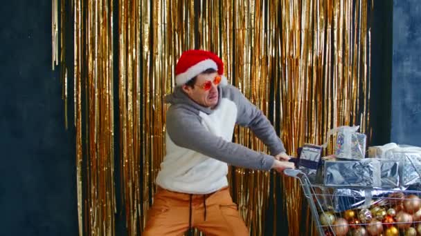 Young male with shopping cart filled of assorted baubles and christmas presents. Man in santa hat and bright glasses pull cart hard with christmas toys and gift boxes. Concept of discounts and sales. — Stock Video
