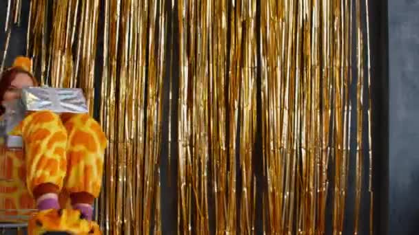 Lächeln im kigurumi mit Weihnachtsgeschenken im Einkaufswagen. Seitenansicht einer aufgeregten jungen Frau im kigurumi-Pyjama, die im Einkaufswagen mit Weihnachtsgeschenken in glänzender Verpackung mit goldenem Lametta an der Wand sitzt — Stockvideo