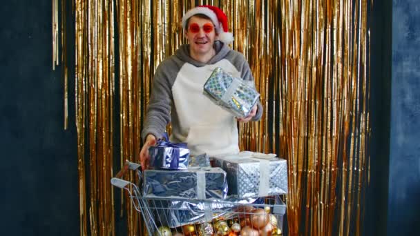 Młody mężczyzna w jasnych okularach i kapelusz Mikołaja stojący z wózkiem wypełnionym różnorodnymi bombkami i zapakowanymi prezentami świątecznymi na ścianę z błyszczącym świecidełkiem. — Wideo stockowe