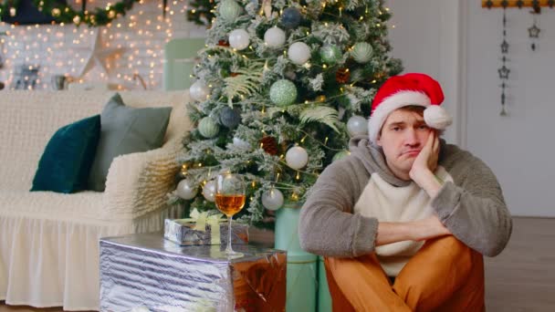 Λυπηρός άντρας με καπέλο Σάντα κάθεται στο πάτωμα κοντά στο χριστουγεννιάτικο δέντρο στο σαλόνι. Μοναχικός άνδρας αναστενάζει, κοιτάζοντας την κάμερα και μετά κάτω κατά τη διάρκεια των διακοπών. — Αρχείο Βίντεο