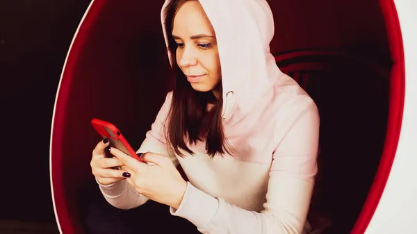 Jovem com telefone celular sentado em cadeira de bola no fundo preto. Feminino smartphone de navegação e descansando em poltrona moderna. — Fotografia de Stock