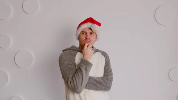 Homem de chapéu de Papai Noel coçando queixo no fundo da parede branca modelada. Macho pensativo olhando para longe e refletindo sobre as próximas férias. — Fotografia de Stock