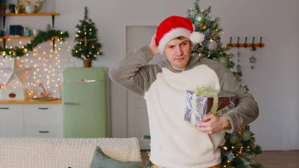 Tänksam hane i Santa hatt klia huvudet när du står med insvept presentförpackning i platt med julgran och glödande girlanger och tittar bort med tvivel blick — Stockfoto