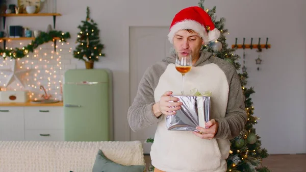 Człowiek z darem i szklanką alkoholu. Mężczyzna w Santa kapelusz stojący w mieszkaniu ozdobiony świąteczną choinką i próbuje pić z kieliszka do wina, stojąc na pudełku prezentów. Człowiek świętuje Nowy Rok. — Zdjęcie stockowe