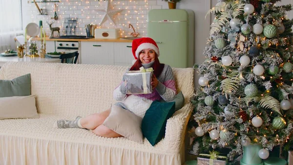 Ung kvinna i tomte hatt med presentask sitter på soffa nära julgran i vardagsrummet. Glad kvinna anser och kramar julklapp. Begreppet överraskning under semestern. — Stockfoto
