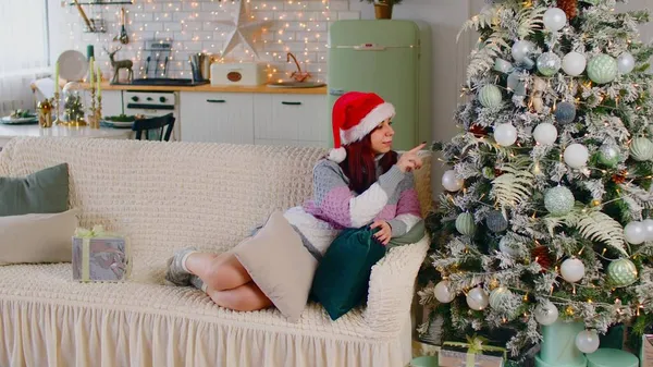 Jeune femme assise sur le canapé et regardant l'arbre de Noël. Femme rêveuse considère et touche décorations de Noël sur l'arbre de fête, assis dans un studio décoré avec des guirlandes rougeoyantes. — Photo