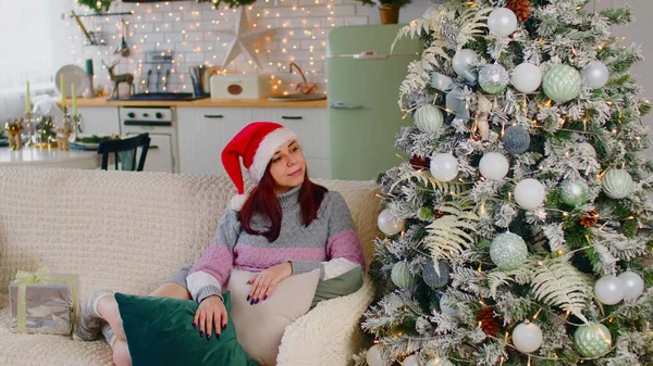 Kvinna beundrar julgran. En vacker brunett i en nyårshatt som beundrar julgranen. Ung hona tittar på en vackert dekorerad julgran — Stockfoto