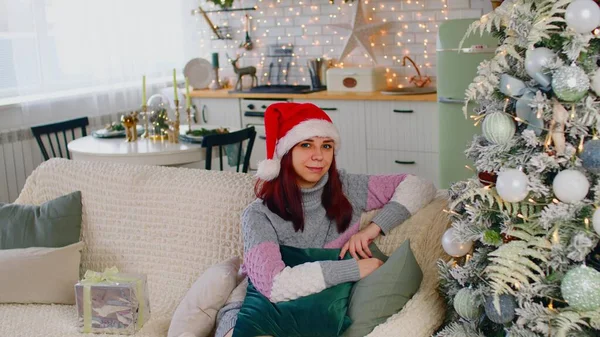 Mooie vrouw in een nieuwjaarshoed zittend op de bank naast een kerstboom en kijkend naar camera — Stockfoto